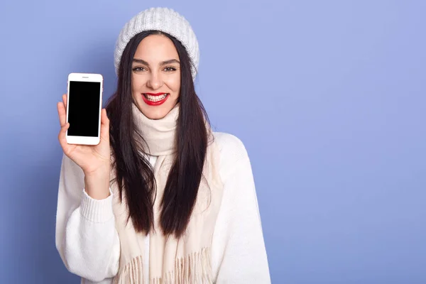 Молодая женщина показывает дисплей мобильного телефона с пустым экраном, стоит улыбаясь изолированы на синем фоне, дама в теплой одежде, копия пространства для рекламы или рекламного текста . — стоковое фото