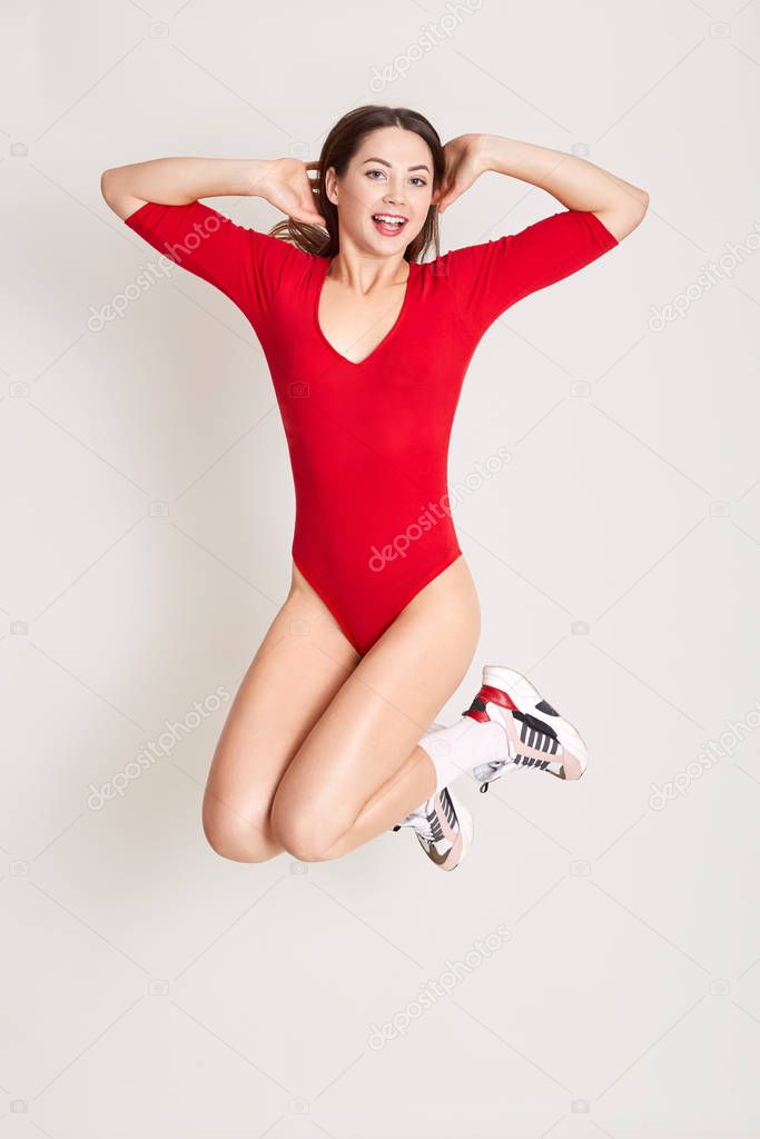 Full length studio photo of attractive woman jumping in air with arms on her head, posing isolated over white background, keeping mouth opened, having excited facial expression, looks happy.