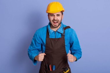 İnşaatçı sarı kask, gömlek ve önlük giyer, tamirat yapar, cebinde aletler taşır, mavi arka planda izole olur. İşçi ya da tamirci çalışmakla meşgul, mutlu bir şekilde bağırıyor..