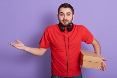 Beyaz sakallı bir adamın stüdyo fotoğrafı. Kırmızı bir tişört giymiş. Elinde karton kutuyla eflatun arka planda duruyor..