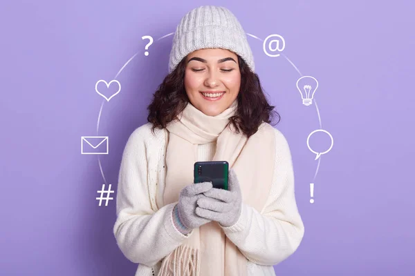 Portrait of happy dark haired girl posing isolated over lilac background in studio, holding smartphone in hands, chatting with pleasure with her boyfriend, thinks what ti type, having toothy smile. — Stock Photo, Image