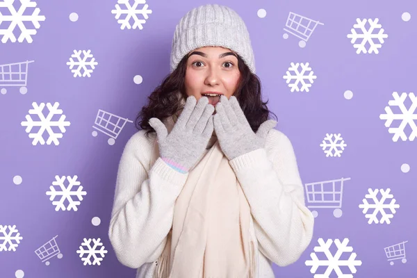 Крупный план портрета девушки в тёплой зимней одежде, вязаном свитере, шляпе и варежках, с удивительным выражением лица, привлекательной женщиной, держащей руки на открытом рте, выглядит удивлённым — стоковое фото
