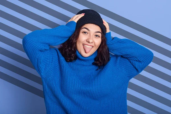 Retrato de chica encantadora con el pelo oscuro, mostrando su lengua y manteniendo las manos en la cabeza, mirando a la cámara aislada sobre fondo azul con rayas diagonales, mujer usa suéter azul y gorra negra . —  Fotos de Stock