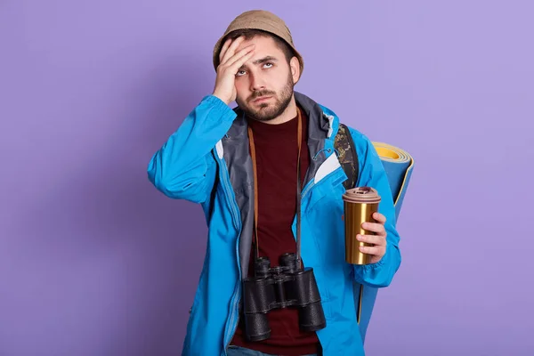 Μπροστά όψη του δυστυχισμένου άνδρα με σακίδιο στο μπλε φόντο, κρατώντας θερμο κούπα με καφέ ή τσάι, κρατώντας το χέρι στο μέτωπο, έχοντας πονοκέφαλο, ξεχνώντας λασπολογία σημαντική. Έννοια ταξιδιού. — Φωτογραφία Αρχείου