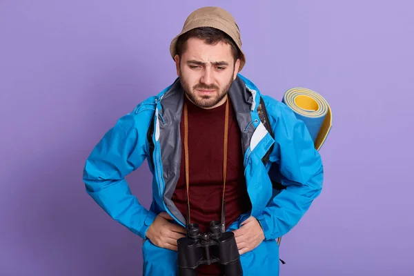 Στούντιο πλάνο του νεαρού όμορφου άνδρα στέκεται απομονωμένη overblue φόντο με τα χέρια στο στομάχι, επειδή δυσπεψία, ταξιδιώτης με χαλάκι και κιάλια, φορώντας σακάκι και καπέλο. Έννοια υγιεινής φροντίδας. — Φωτογραφία Αρχείου