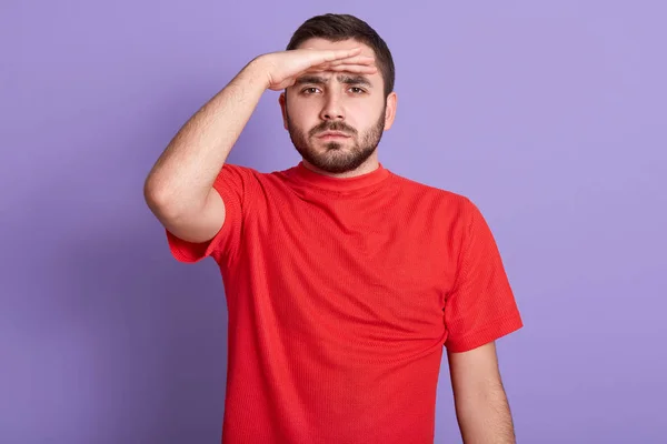 Nahaufnahme Porträt eines ernst zu nehmenden bärtigen Mannes, der in die Ferne blickt, die Hand an der Stirn hält, isoliert vor blauem Hintergrund posiert, attraktiver Typ in rotem lässigem T-Shirt. Menschen-Konzept — Stockfoto