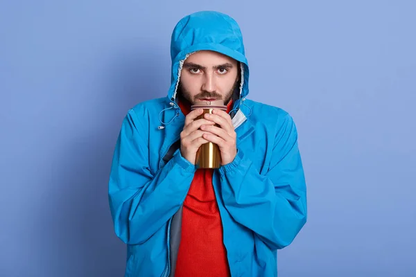 Στιγμιότυπο από τον ελκυστικό νεαρό άνδρα με γενειάδα, με δυστυχισμένη έκφραση προσώπου, κρατώντας θερμό ποτήρι με καφέ ή τσάι, ντύνεται μπλε σακάκι με κουκούλα και κόκκινο πουκάμισο, όντας ψυχρός, καλλωπιστικά χέρια. — Φωτογραφία Αρχείου