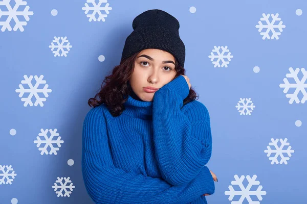 Close up retrato de jovem morena do sexo feminino em pé sobre a parede do estúdio com flocos de neve no fundo, mantenha a mão sob o queixo e perca alguém, olhando chateado, vestindo camisola azul e boné preto . — Fotografia de Stock