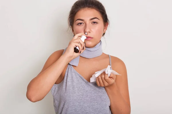 生病的女人拿着擦拭的手，用鼻腔喷雾器，在白色的背景上摆出孤立的姿势，穿着休闲装，感觉不舒服，女孩面带微笑地看着相机. — 图库照片