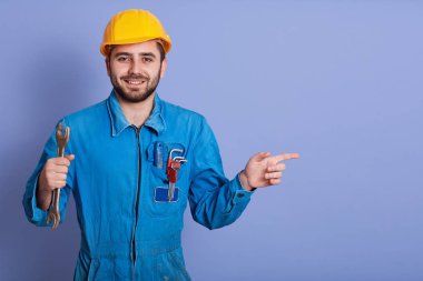 Elinde İngiliz anahtarı olan mutlu ve gülümseyen sakallı mühendis kameraya bakıyor ve mavi stüdyo arka planına karşı dururken işaret parmağıyla işaret ediyor, mavi üniformalı ve sarı kasklı..