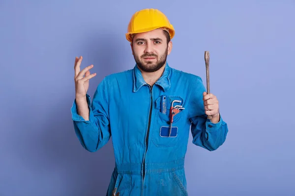 Zmatený samec instalatér nebo automechanik nechápe, kde je zlomenina, pokrčí rameny a zvedne ruce v úžasu, nosí žlutou helmu a modrou uniformu, pózuje proti zdi sztudio. — Stock fotografie