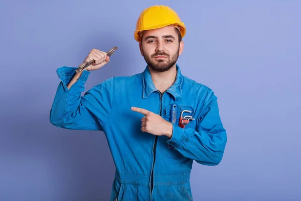Detailní portrét mladého pohledného ručního dělníka držícího v ruce klíč a ukazujícího na své silné biceps izolované přes modré pozadí, automechanik ukazující, jak je silný. Koncept lidí. — Stock fotografie