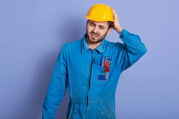 Studio záběr pohledného mladého vousatého bělošského mechanika v modrém pracovním oděvu a žlutém tvrdém klobouku, stojí a dotýká se hlavy, má bolestivý pohled, po náročném pracovním dni se cítí vyčerpaný. — Stock fotografie