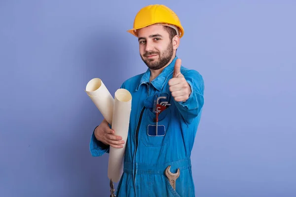 Mladý bělošský architekt s přilbou, pohledný muž pózující s palcem nahoru a držící bluepint v rukách, v modré uniformě a žlutém tvrdém klobouku, pracuje na novém projektu, má velké úspěchy. — Stock fotografie