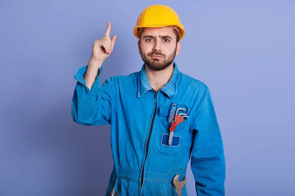 Studio záběr mladého stavebního dělníka s nářadím v kapse, šaty žlutá přilba a uniforma ukazuje nahoru, vousatý muž s vážným výrazem, kopírovací prostor pro reklamu nebo propagační text. — Stock fotografie