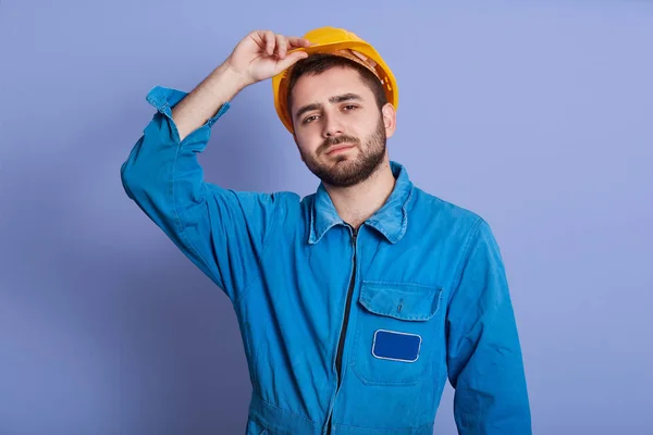 Studiový záběr muže v modré uniformě a žluté přilbě, jak se dívá do kamery a drží ruku na klobouku, pohledného chlapa s tmavými vousy, vypadá unaveně po tvrdé práci. Koncept budovy. — Stock fotografie