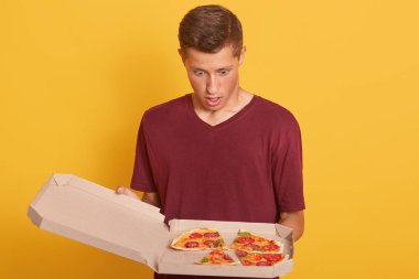 Pizzaya bakan adamın ağzı açık ve şaşkın bir yüz ifadesi olan, sıradan tişört giyen, sarı stüdyo duvarının önünde duran çekici bir adam. Teslimatçı siparişleri karıştırır..
