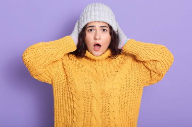 Dalgalı saçlı güzel esmer kadın leylak arka planda tek başına duran sarı kış süveteri giyiyor, deli gibi görünüyor ve elleri başının üstünde korkmuş, korkmuş ve şok olmuş, ağzı açık poz veriyor..