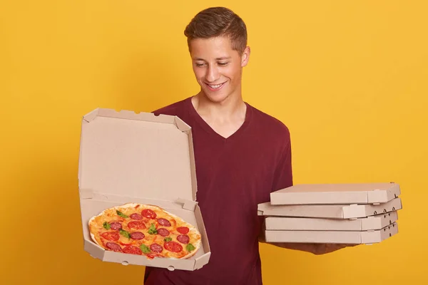 Handsome young delivery worker dresses burgundy casual t shirt holding pizza in boxes, looking at opened box and smiling, attractive male stands against yellow studio wall, looks happy, doing his job. — Stock Photo, Image