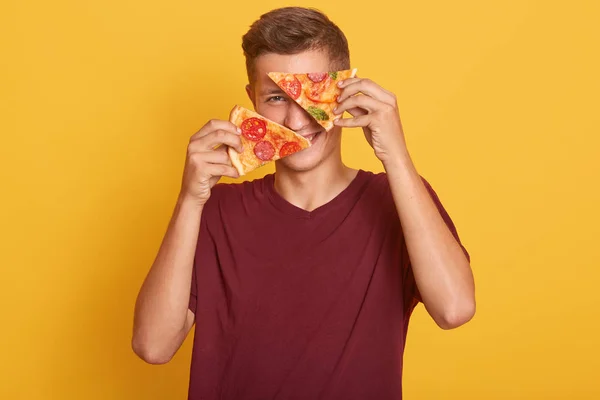 Joven sosteniendo dos piezas de deliciosa pizza en sus manos y cubriéndose los ojos con un producto sabroso, posando aislado sobre el fondo amarillo del estudio, disfrutando de un divertido tiempo libre, listo para comer . — Foto de Stock