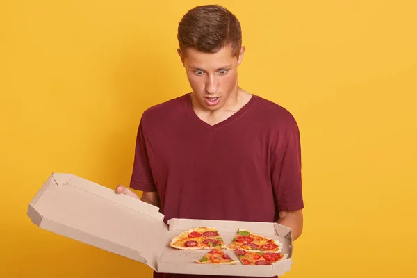 Gros plan de l'homme regardant la pizza avec la bouche ouverte et l'expression faciale étonnante, gars attrayant portant casual chemise bordeaux, debout contre le mur jaune studio. Livraison homme mélange l'ordre . — Photo