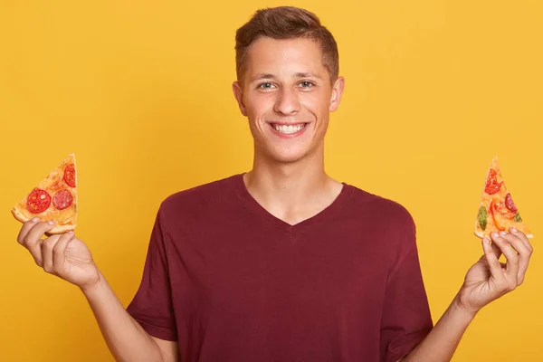 Vrolijke glimlachende man met twee stukken pizza in de hand, klaar voor een heerlijke snack. Happy man beveelt pizza, mannelijk model jurken bordeaux casual t-shirt, poseren geïsoleerd over gele muur — Stockfoto