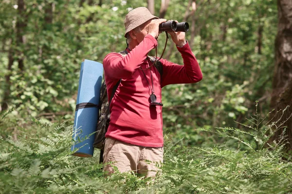 Shiot extérieur de l'homme regardant à travers les jumelles dans la forêt, voyageur âgé portant des vêtements décontractés ayant l'aventure dans le bois, trouver la bonne façon, posuing autour des arbres, a tapis et sac à dos sur son dos . — Photo