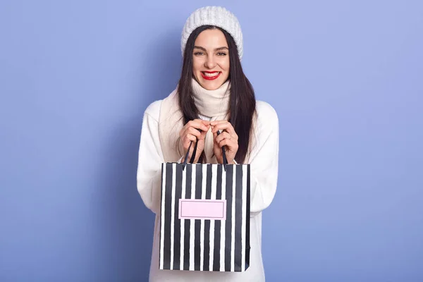 Imagem de senhora positiva enérgica com cabelos pretos e lábios vermelhos, segurando presente no saco listrado de papel, sorrindo sinceramente, olhando diretamente para a câmera, de pé isolado sobre fundo lilás em estúdio . — Fotografia de Stock