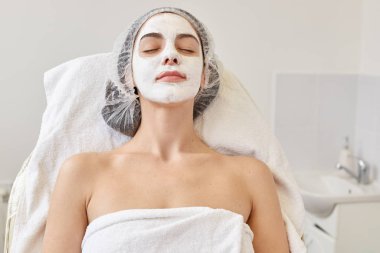 Yüzünde maske olan genç bir kızın portresi beyaz arka planda kapalı gözlerle yatıyordu. Kozmetik prosedür, güzellik kaplıcası ve kozmetik konsepti. Kız salonda dinleniyor..