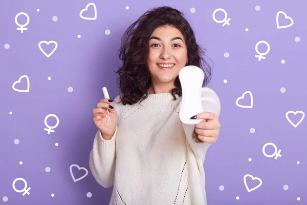 Imagen de una hermosa mujer joven y positiva sosteniendo almohadilla de higiene y tampón en ambas manos, eligiendo almohadilla sanitaria, usando suéter blanco, con expresión facial agradable. Concepto de menstruación . — Foto de Stock