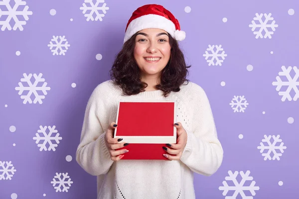 Фото веселой мирной девушки в красной шляпе Санта Клауса и белом свитере, держащей красную коробку в обеих руках, получающей рождественские подарки, стоящей изолированной на фоне сирени в студии . — стоковое фото