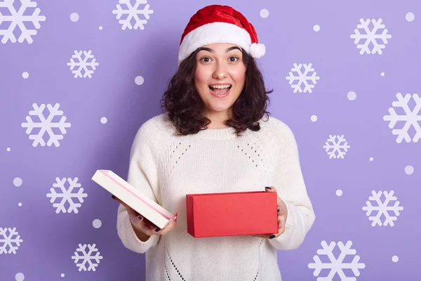 令人惊讶的是，黑发发女郎面容震惊，打开红白相间的盒子，兴高采烈，拿着圣诞礼物，头戴红色的圣诞老人帽，头戴白色毛衣。 休息概念. — 图库照片