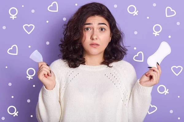 Foto de morena emocional confundida mirando directamente a la cámara, habiendo sorprendido la expresión facial, sosteniendo la copa menstrual y la almohadilla de higiene en ambas manos, aislada sobre fondo lila en el estudio . — Foto de Stock