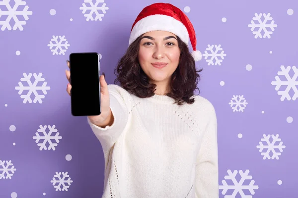 Image of attractive energetic brunette wearing white sweater and red santa claus hat, standing isolated over lilac background with snowdrops, holding smartphone with blank screen in one hand. — Stock Photo, Image