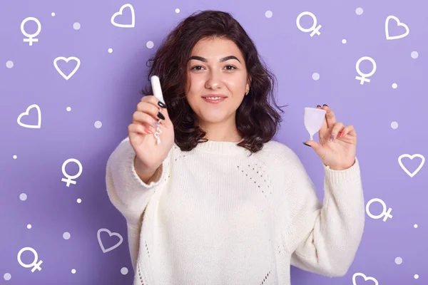 Ritratto di bella donna dai capelli neri positiva che tiene tampone e assorbente igienici, guarda direttamente la fotocamera, usando tampone durante i periodi, in piedi isolato sopra il backgound lilla in studio . — Foto Stock