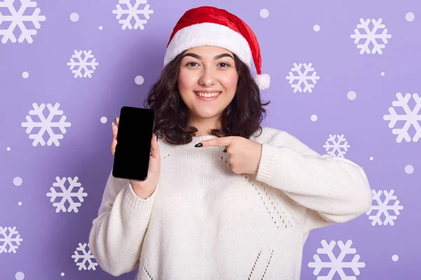 Портрет крупным планом улыбающейся темноволосой женщины, указывающей пальцем на смартфон, держащей мобильный телефон в одной руке, смотрящей прямо в камеру. Современная концепция альтернатив . — стоковое фото