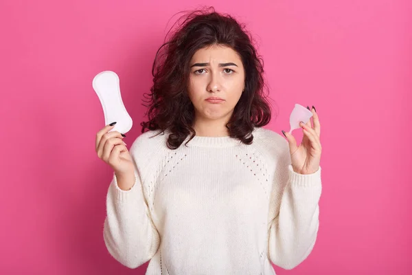 Tiro de estudio interior de linda mujer molesta insatisfecha sosteniendo almohadilla de higiene y copa menstrual, usando suéter blanco, estar de mal humor, tener días críticos, tomar decisiones. Concepto de días críticos . — Foto de Stock