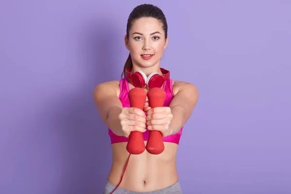 Foto de alegre mujer joven positiva sosteniendo pesas rojas frente a sí misma, teniendo entrenamiento, manteniendo un estilo de vida saludable, disfrutando del deporte, siendo aficionada a las actividades. Concepto Fitness . — Foto de Stock