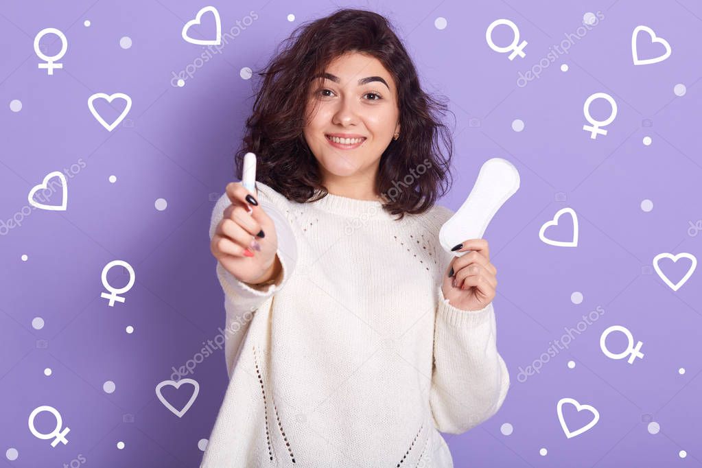 Portrait of cheerful good looking female holding tampon on foreground, having hygiene pad in one hand, looking directly at camera, wearing white sweater, making choice. Critical days concept.