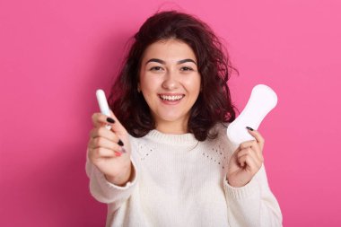 Adet döngüsü sırasında elinde farklı tipte kadın hijyen ürünleri tutan mutlu beyaz kadın. Fwemales 'in ellerinde hijyenik ped ve pamuk tampon. Pembe arka planda tek başına duruyor..