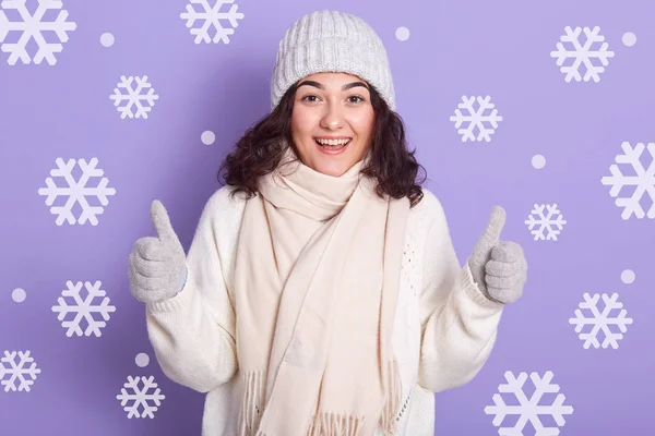 Зимняя девушка с темными волнистыми волосами, одетые в белый теплый свитер, шарф, кепку и варежки, стоящие с большими пальцами вверх изолированы над стеной студии со снежинкой на заднем плане, девушка выглядит счастливой и взволнованной . — стоковое фото