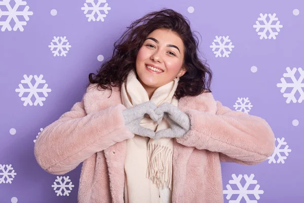 Ritratto ravvicinato di giovane donna formata cuore con le mani, attraente ragazza dai capelli scuri indossa cappotto di eco pelliccia rosa pallido, sciarpa e guanti, posa isolata sulla parete lilla con fiocchi di neve sullo sfondo . — Foto Stock