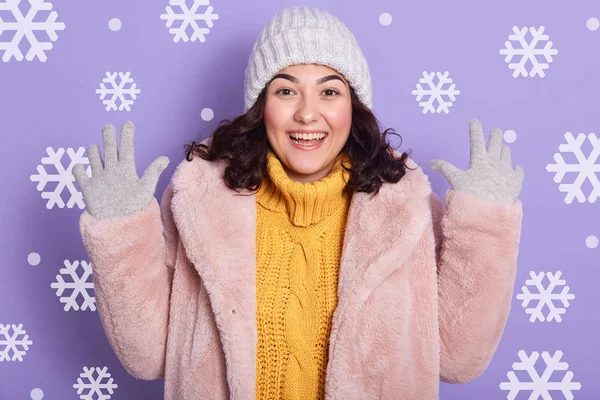 Изображение женщины в стильной эко-шубе, желтом свитере и кепке, смотрящей в камеру и держащей руки в студии со снежинками на заднем плане. Женщина выглядит взволнованной и выражает счастье . — стоковое фото