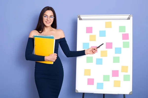 Attractive smiling Caucasian woman in office next to whiteboard with multicolored stickers memos, girl with bare shoulders pointing at her scheme, female wearing dresses and glasses, holds folder. — Stock Photo, Image