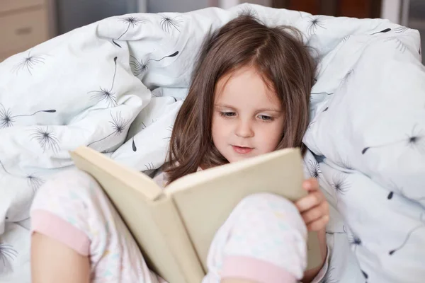 Close up indoor shot of little cute girl dresses pajama reading interesting book while lying in bed, darkhaired female child looks concentrated, relaxing in morning, enjoying stories or fairytailes. — Stock Photo, Image