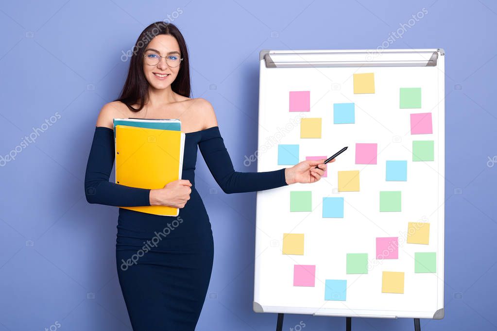 Attractive smiling Caucasian woman in office next to whiteboard with multicolored stickers memos, girl with bare shoulders pointing at her scheme, female wearing dresses and glasses, holds folder.