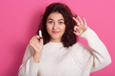 Siyah kıvırcık saçlı, jest yapan, bir elinde tampon tutan, doğrudan kameraya bakan, adet gören pozitif görünümlü genç bir kızın yatay çekimleri. Regl konsepti.