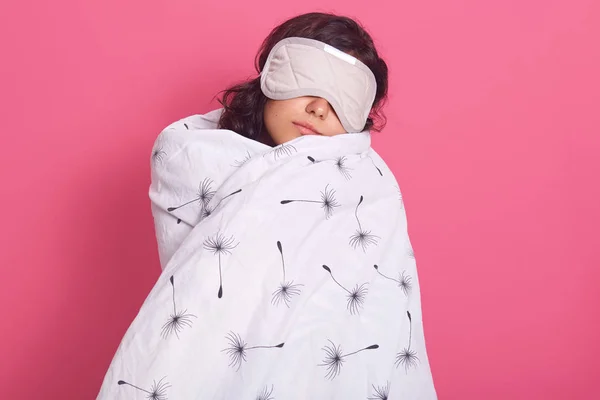 Sleep equipment concept. Portrait of brunette woman wrapped white blanket and wearing sleeping eye mask. Studio shot of young female isolated over pink background, lady being ready to fall asleep. — Stock Photo, Image