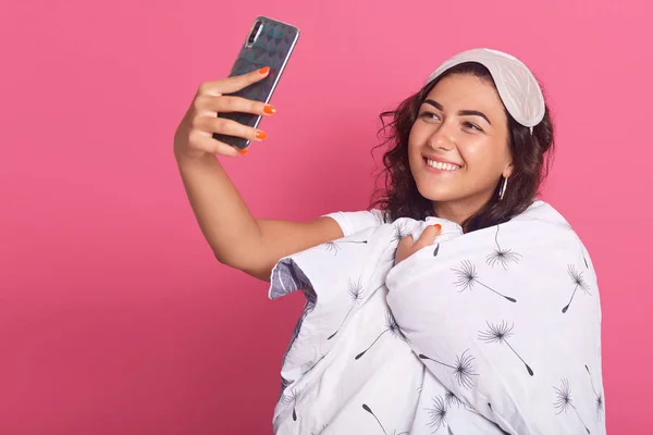 Foto de estudio de una joven morena con máscara de dormir y manta blanca envuelta, tomando selfie durante la hora de acostarse, chica divirtiéndose en casa por la mañana antes de ir a trabajar, aislada sobre el concepto rosa . — Foto de Stock