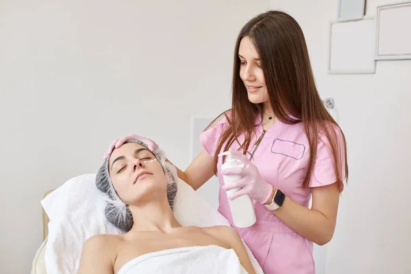 Image of cute professional dermatologist doing skin treatment, improving skin condition, holding special facial gel, applying it to skin, female lying in beauty parlor, closing eyes, getting pleasure.